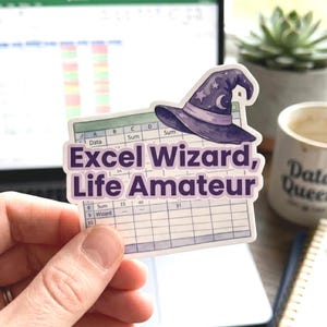 May include: A die-cut sticker featuring a purple wizard hat with stars and a crescent moon resting atop a spreadsheet grid. The text "Excel Wizard, Life Amateur" is printed in bold purple letters across the center of the spreadsheet design.