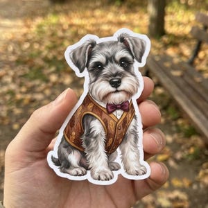 May include: A die-cut sticker featuring a gray and white Schnauzer dog sitting upright. The dog wears a brown vest patterned with autumn leaves and a small dark red bow tie. The illustration has a white border and a smooth, matte finish.