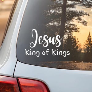 May include: White decal on a car window with the words "Jesus King of Kings" in a script font. The background shows a blurred view of trees and a sunset.