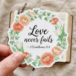 May include: A white sticker with the text "Love never fails - 1 Corinthians 13:8" in black script, surrounded by a floral wreath of peach roses, white tulips, and green ivy. The sticker is held in a hand.