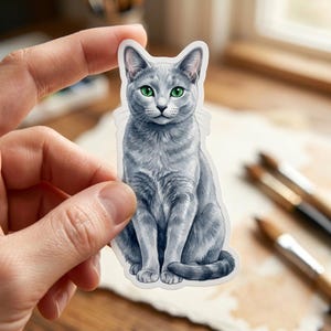 May include: A sticker featuring a seated gray cat with bright green eyes. The cat has detailed fur and is held between two fingers. The background includes paintbrushes and a watercolor painting.