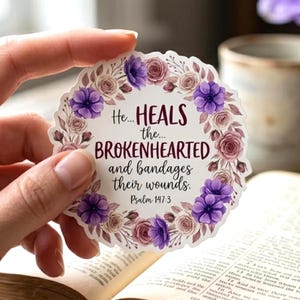 May include: A circular sticker featuring a floral wreath of purple anemones and dusty pink roses surrounding the text: He... HEALS the... BROKENHEARTED and bandages their wounds. Psalm 147:3. The design is printed on a white background with scalloped edges.