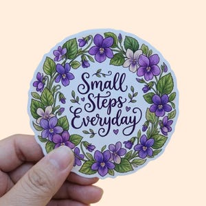 May include: A round sticker with a floral border of purple violets and green leaves. The sticker has the words "Small Steps Everyday" in a decorative script font.