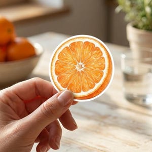 May include: A circular sticker featuring a detailed illustration of a sliced orange. The design displays the vibrant orange pulp, segmented sections, and a white rind edge, capturing the natural texture and bright citrus colors of the fruit.