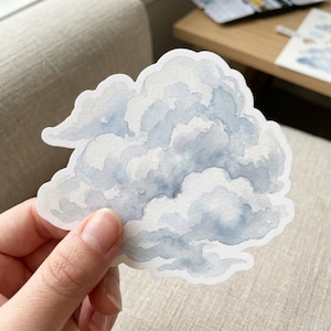 May include: A die-cut sticker featuring a watercolor painting of a cloud. The cloud is primarily light blue and white, with soft edges and a fluffy appearance. The sticker is held by a hand against a neutral background.