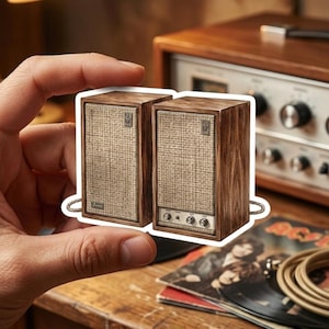 May include: A sticker featuring two rectangular speakers with a dark wood grain finish and beige fabric grilles. The right speaker includes three silver control knobs and a small silver logo plate at the top corner of the grille on both units.