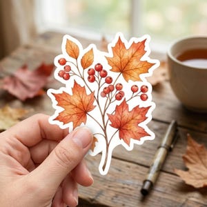 May include: A die-cut sticker featuring a watercolor illustration of autumn foliage, specifically orange and red maple leaves mixed with clusters of small red berries on thin brown stems, all outlined with a clean white border.