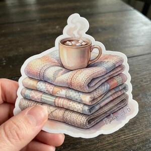 May include: A sticker featuring a stack of folded blankets with a mug of hot chocolate on top. The blankets are in shades of pink, blue, and beige. The mug has marshmallows and steam rising from it. The text "Psalm 19:76" is on the sticker.
