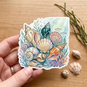May include: Watercolor sticker featuring a collection of seashells in various shapes and colors, including tan, orange, and teal. The design includes coral, seaweed, and small fish, creating an underwater scene.