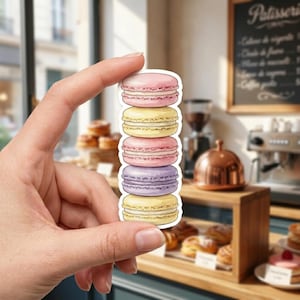 May include: A die-cut sticker featuring a vertical stack of five colorful macarons. The stack consists of a pink macaron on top, followed by yellow, pink, purple, and yellow macarons at the bottom. Each macaron has a visible cream filling.