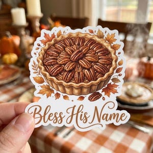 May include: A die-cut sticker featuring a pecan pie topped with brown nuts, surrounded by orange and brown autumn leaves. Below the pie, the text Bless His Name is written in a brown cursive script on a white background.