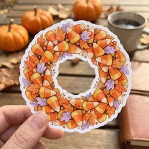 May include: A circular sticker featuring a wreath design composed of orange, yellow, and white candy corn pieces interspersed with small orange and light purple autumn leaves, tiny berries, and small star shapes on a white background.