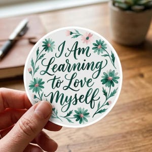 May include: A circular sticker with a white background featuring the dark green cursive text I Am Learning to Love Myself. The text is surrounded by a decorative wreath of watercolor-style teal green daisies and small pink flower buds.