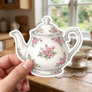 May include: A die-cut sticker shaped like a white porcelain teapot featuring a delicate pink rose floral pattern with green leaves. The teapot has a matching lid, a curved spout, and a handle, all decorated with the same floral motif.