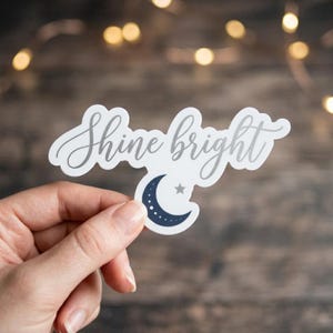 May include: A die-cut vinyl sticker featuring the text Shine bright in a grey cursive script font. Below the text is a dark navy blue crescent moon graphic with small white dots along its curve and a small grey star positioned above the moon.
