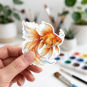 May include: A sticker featuring a detailed illustration of a goldfish with flowing orange fins and tail. The fish has a golden body with scales and is surrounded by small bubbles and watercolor accents. The sticker is held by a hand.