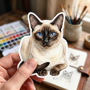 May include: A sticker featuring a watercolor illustration of a Siamese cat. The cat has striking blue eyes, a dark brown face, ears, and paws, and a cream-colored body. The sticker is held by a hand.
