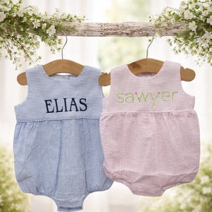 Könnte beinhalten: Zwei Baby-Strampler hängen an Holzbügeln. Einer ist hellblau mit vertikalen Streifen und dem Namen "ELIAS" in Marineblau gestickt. Der andere ist rosa mit einem gepunkteten Muster und dem Namen "sawyer" in Grün gestickt.