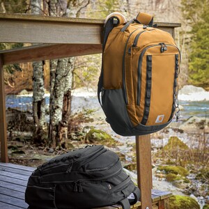 Puede incluir: Dos mochilas, una marrón y otra negra, sobre una terraza de madera. La mochila marrón cuelga de un poste de madera. Las mochilas tienen múltiples compartimentos y correas.