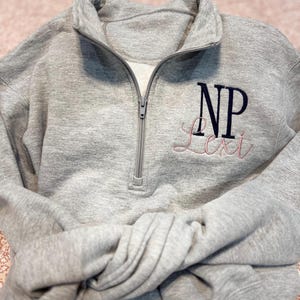 May include: Gray quarter-zip sweatshirt with a navy blue monogram "NP" and the name "Lexi" embroidered in pink.
