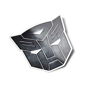 Op de afbeelding: Een zwarte en grijze metalen sticker met het Transformers-logo. Het logo is een gestileerd robotgezicht met een geometrisch ontwerp.