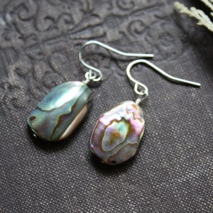 Pode incluir: Um par de brincos iridescentes de concha de abalone com ganchos de orelha em prata esterlina. As conchas em forma de gota exibem uma mistura de tons verdes, azuis, rosa e roxos. Um acessório único.