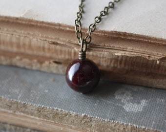 May include: A dark red, round gemstone pendant necklace with an antique brass chain. The pendant is a polished, spherical shape. The necklace rests on a stack of old, worn books with aged pages.