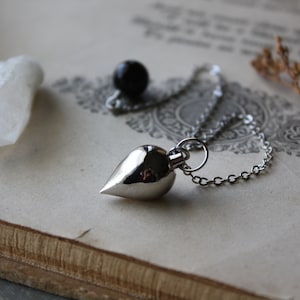May include: A silver pendulum necklace with a teardrop-shaped pendant and chain. A small black sphere is near the pendulum. The items are on a vintage book with a decorative pattern. A white crystal and dried plant stems are also present.