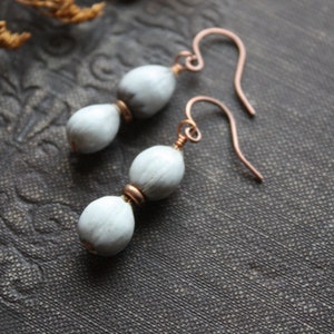 May include: A pair of handmade earrings featuring light gray, oval-shaped beads and copper-colored wire hooks. The earrings have a simple, elegant design, perfect for everyday wear. The beads are connected with small copper-colored rings.