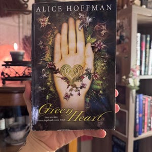 May include: A paperback book titled Green Heart by Alice Hoffman, featuring a cover illustration of an open palm with a gold heart symbol at the centre, surrounded by dark foliage, purple flowers, and the text Green Angel and Green Witch.