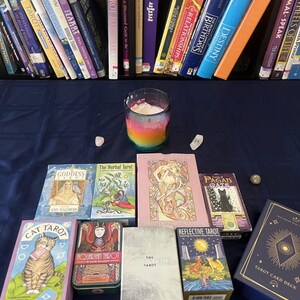 May include: A collection of tarot card decks and books, including "Cat Tarot," "The Herbal Tarot," and "Tarot Card Deck." A rainbow-colored candle and crystals are also present, all arranged on a dark blue surface.