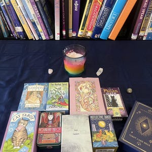 May include: A selection of tarot card decks and books, such as "Cat Tarot," "The Herbal Tarot," and "Tarot Card Deck." A rainbow-hued candle and crystals are also visible. The items are displayed on a dark blue surface.