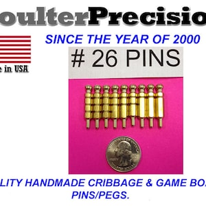 Peut inclure: Un ensemble de 26 pions de cribbage en métal couleur or, disposés en rangée sur une surface rose. Les pions ont un sommet arrondi et un fond pointu. L'image comprend également un petit drapeau américain et le texte "Coulter Precision" et "Since the year of 2000".