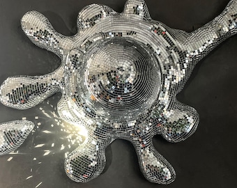 May include: A large, silver, disco ball-style sculpture in a unique, organic shape, resembling a splash or amoeba. Several smaller, similar shapes are scattered around it, all reflecting light. The background is a dark, matte surface.