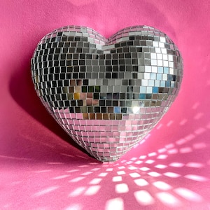 May include: A heart-shaped disco ball with a mirrored surface. The ball is made up of many small, square mirrored tiles. The heart is set against a pink background, with the light reflecting off the mirrored surface.