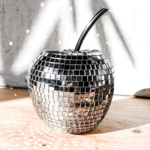 May include: A decorative silver apple sculpture with a mirrored disco ball effect. The apple has a shiny, reflective surface made of small square tiles. A curved, black stem extends from the top. The sculpture is placed on a wooden surface.
