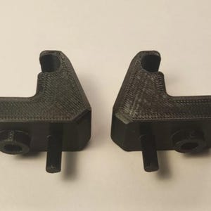 Puede incluir: Dos componentes de plástico negro impresos en 3D. Cada uno tiene una base rectangular con una proyección cilíndrica y un poste vertical. La sección superior tiene una forma curva, similar a un gancho. Los artículos están sobre un fondo liso.