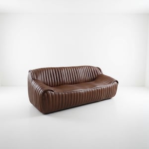 May include: A brown leather sofa with a ribbed design. The sofa has a rounded shape and is set against a white background. The sofa appears to be made of high-quality leather.