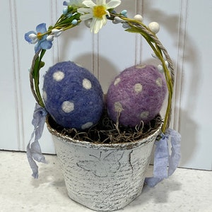 May include: A white ceramic planter with a floral wreath handle, filled with brown straw and two felt Easter eggs. One egg is purple with white polka dots, and the other is blue with white polka dots.
