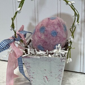 May include: A small white painted basket with a pink and blue polka dot felt egg inside. The basket is decorated with a white vine with small white flowers and a pink and blue gingham ribbon.