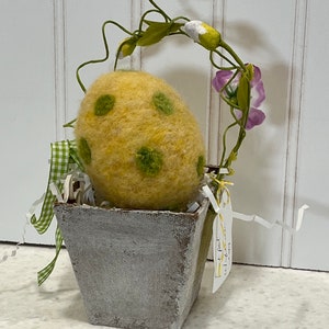 May include: A yellow felt Easter egg with green polka dots sits in a gray planter with a green and white gingham ribbon. A vine with pink and white flowers arches over the egg.