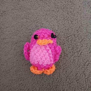 May include: A pink rubber band bird with orange feet and black eyes. The bird is made with a loom.