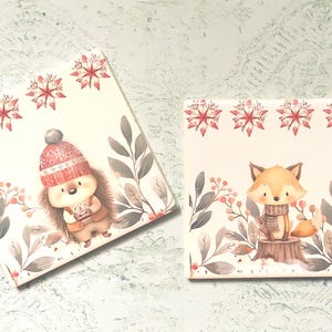 Könnte beinhalten: Zwei quadratische Keramikuntersetzer mit Illustrationen von Waldtieren. Ein Untersetzer zeigt einen Igel mit roter Strickmütze, der andere einen Fuchs mit Schal. Beide haben rote Sternmuster am oberen Rand und graue Blattmotive.