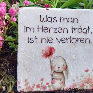 Könnte beinhalten: Eine quadratische Steinplatte mit dem deutschen Text "Was man im Herzen trägt, ist nie verloren." Die Platte zeigt eine aufgedruckte Illustration eines kleinen Hasen, der eine rosa Blume hält, umgeben von kleinen rosa Blumen am unteren Rand.