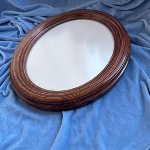 May include: An oval-shaped mirror featuring a polished, dark brown wooden frame with a visible natural grain pattern. The frame has a multi-layered, stepped edge design that creates a sense of depth around the reflective glass surface.