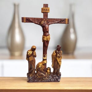 Könnte beinhalten: Eine Holzfigur, die die Kreuzigung Jesu Christi mit zwei knienden Figuren an der Basis darstellt. Das Kreuz ist braun mit goldenen Akzenten und die Figuren sind in Braun- und Goldtönen bemalt. Die Inschrift "I.N.R.I." ist auf dem Kreuz zu sehen.