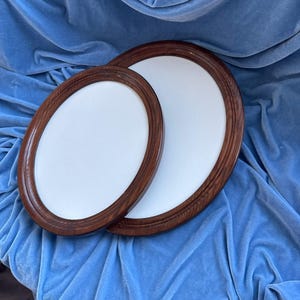 Könnte beinhalten: Zwei ovale Bilderrahmen mit einer dunkelbraunen Holzmaserung und glatten, weißen, leeren Zentren. Die Rahmen sind überlappend angeordnet und weisen ein abgerundetes, geriffeltes Kantendesign entlang des gesamten Umfangs jedes Rahmens auf.