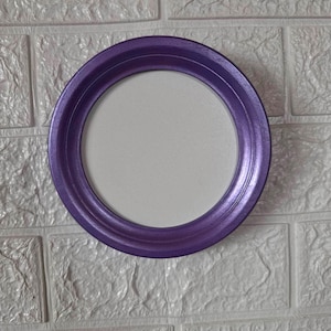 May include: A circular frame with a smooth, metallic purple finish surrounding a plain, light-colored circular center. The frame features a rounded, raised edge that creates a distinct border around the inner white circular surface.