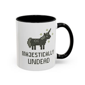 May include: White ceramic mug with a black rim and handle. The mug features a pixelated black unicorn with a horn and a green sparkle effect. The text "MAJESTICALLY UNDEAD" is printed in black below the unicorn.