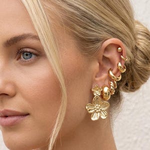 Könnte beinhalten: Nahaufnahme eines Ohrs, das mit mehreren goldenen Ohrringen geschmückt ist. Die Ohrringe umfassen kleine Ohrstecker, Creolen und ein großes blütenförmiges Design. Der Schmuck hat eine helle, glänzende Goldfarbe.
